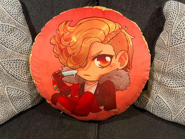 A stuffed round pillow of Ankh from Kamen Rider OOO. Drawn in a chibi style, he is holding a melting popsicle to his face and scowling. The background is the Taka medal.