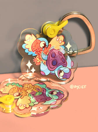 A photo showing two keychains of Archen and Tirtouga with a heart-shaped clasp. The upright charm shows their normal colours while the charm laying flat on the table is their shiny colours.